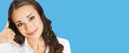 Businesswoman in white confident clothing showing call me hand sign gesture, isolated on bright blue color background. Portrait of smiling gesturing brunette woman at studio. Business concept photo.の写真素材