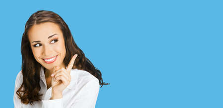 Young happy smiling woman in white confident blouse, showing pointing at copy space for text or imaginary. Business ad concept. Isolated on blue color background. Brunette businesswoman. Wide.の写真素材