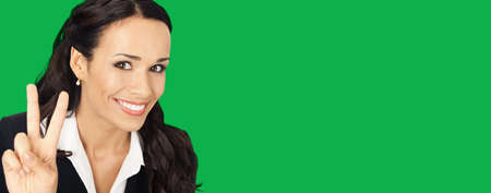 Portrait image of businesswoman in black confident suit showing two fingers or victory gesture, isolated on vivid green background. Happy smiling woman at studio. Business success concept.の写真素材