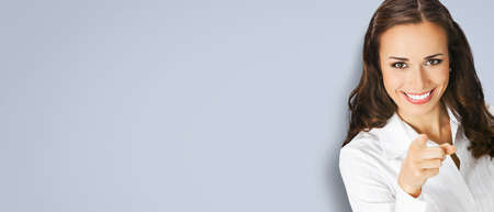 What about You? Happy smiling young woman in white confident blouse pointing finger at viewer, copy space for some text. Business concept. Grey color background. Brunette businesswoman. Wide.の写真素材