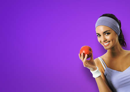 Portrait image of african american woman in sportswear with red apple, isolated on violet purple background. Young sporty model at studio. Dieting, fitness, dental care ad concept.の写真素材
