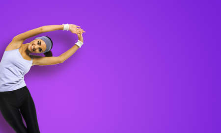 Happy smiling african american woman in sportswear doing stretching fitness exercise or yoga moves, isolated on violet purple background. Sporty girl at studio. Copy space empty text areaの写真素材