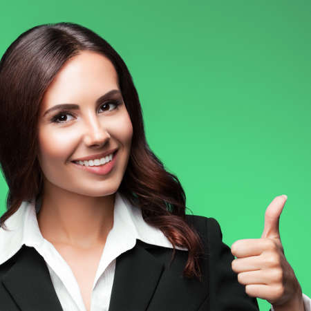 Portrait image of happy smiling young businesswoman in black suit, showing thumb up gesture, isolated on green background. Square composition.の写真素材