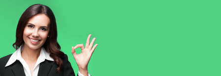 Portrait of happy smiling young business woman in black suit, showing okay gesture, with copy space for some slogan or text, isolated on green background.の写真素材