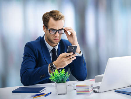 Serious businessman in glasses and blue suit working with cell smart phone and laptop at office. Success in business education concept image.の写真素材