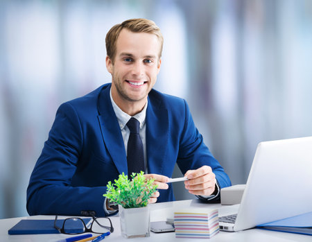 Young happy smiling businessman in blue suit working with laptop at office. Success in business, job concept image.の写真素材