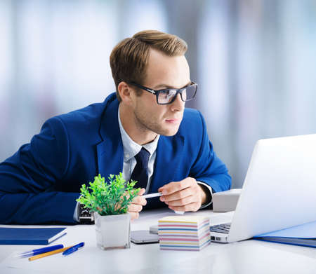 Thoughtful businessman in glasses and blue suit working with laptop computer at office. Business, job and education concept image.の写真素材