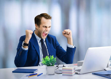 Very happy business man in confident suit, working with laptop at office. Excited business man rising hands up with clenching fists - success in job concept image.の写真素材
