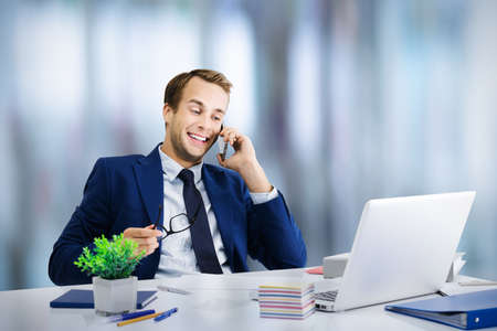 Young happy smiling business man and blue suit working with cellphone and laptop computer. Cheerful businessman at office workplace.の写真素材