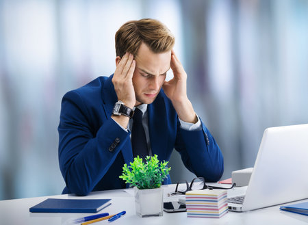 Stressed, tired businessman or heaving headache, holding his head, in blue suit, working with laptop at office. Business fall and education concept image.の写真素材