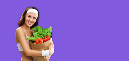 Happy smiling brunette woman in fitness wear holding grocery shopping bag with vegetarian raw food, isolated on violet purple color studio background. Healthy eating and dieting concept.の写真素材