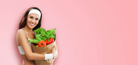 Portrait image of happy smiling brunette woman in fitness wear holding grocery shopping bag with vegetarian raw food, vegetables, isolated on rose pink color background. Healthy eating dieting conceptの写真素材
