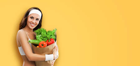 Portrait image of happy smiling young brunette woman in fitness wear holding grocery shopping bag with vegetarian raw food, vegetables, isolated on yellow background. Healthy eating, dieting concept.の写真素材