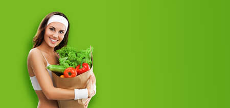 Happy smiling young beautiful brunette woman in fitness wear holding grocery shopping bag with vegetarian raw food, isolated on green color studio background. Healthy eating and dieting concept.の写真素材