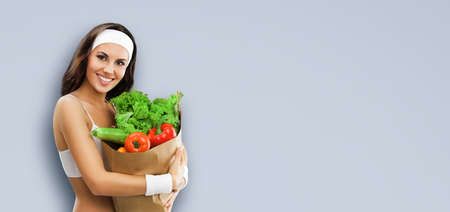 Portrait image of happy smiling beautiful brunette woman in fitness wear holding grocery shopping bag with vegetarian raw food, vegetables, isolated on grey background. Healthy eating, dieting conceptの写真素材