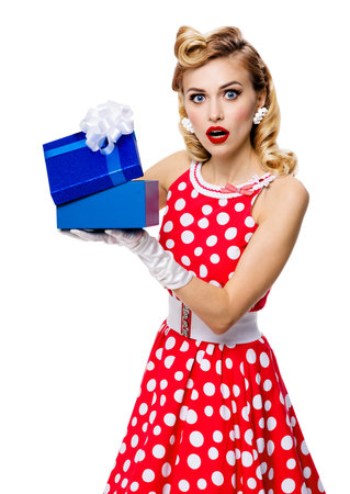 Image of beautiful woman dressed in pin up red dress in polka dot white gloves opening gift box, isolated on white background. Blond pinup model in retro fashion studio concept.の写真素材