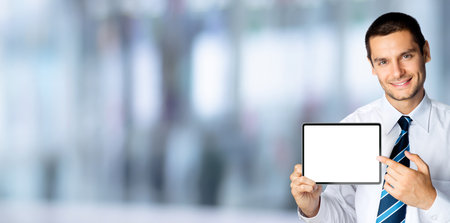 Happy smiling businessman in white shirt and tie, showing pointing tablet pc, touchpad, with copy space, over blurred office interior background. Confident business man.の写真素材