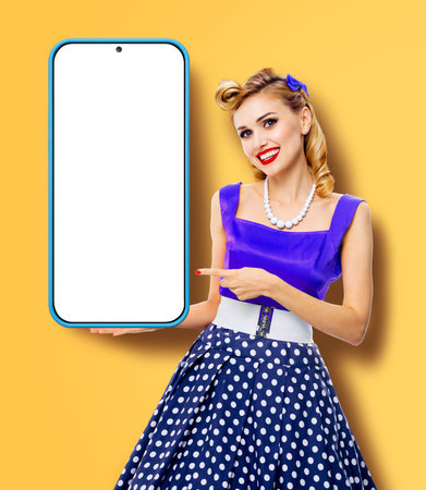 Beautiful smiling woman in blue pinup dress holding showing big smartphone cellphone with mock up screen. Pin up girl advertising, isolated on yellow background. Mobile cell phone app ad concept imageの写真素材