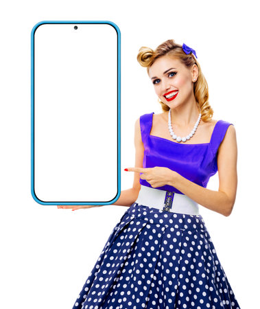 Beautiful smiling woman in blue pinup dress holding, showing big smartphone cellphone with mock up screen. Pin up girl advertising, isolated on white background. Mobile cell phone app ad concept imageの写真素材