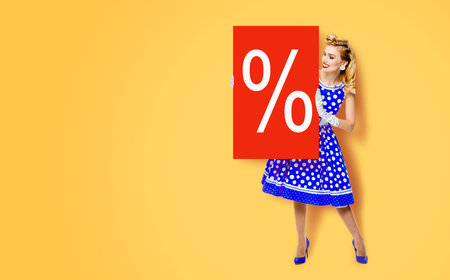 Beautiful woman in pinup dress holding, showing red board with procents % sign. Full body pin up girl with advertising offer, isolated on yellow color background. Sales, discounts rebates ad concept.の写真素材