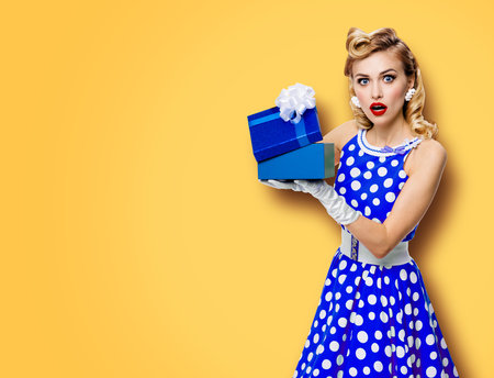 Image of beautiful woman dressed in pin up blue dress in polka dot white gloves opening gift box, isolated on yellow color background. Blond pinup model in retro fashion studio concept.の写真素材