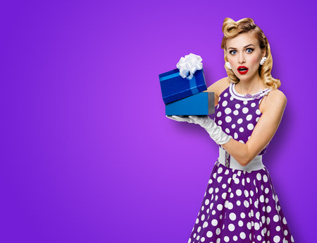 Image of beautiful woman dressed in pin up dress with polka dot white gloves opening gift box, isolated on violet purple background. Blond pinup model in retro fashion studio concept.の写真素材