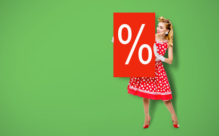 Beautiful woman in pinup dress holding, showing red board with procents % sign. Full body pin up girl with advertising offer, isolated over green background. Sales, discounts rebates ad concept.の写真素材