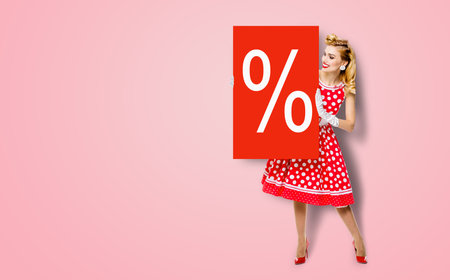 Beautiful woman in pinup dress holding, showing red board with procents % sign. Full body pin up girl with advertising offer, isolated on rose pink background. Sales, discounts rebates ad concept.の写真素材