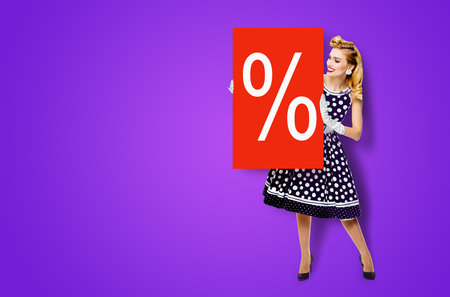 Beautiful woman in pinup dress holding, showing red board with procents % sign. Full body pin up girl with advertising offer, isolated on violet purple background. Sales, discounts rebates ad concept.の写真素材