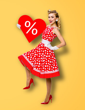 Woman holding red paper heart shape with % sign. Full length body portrait of happy pin up girl. Blond model at retro fashion rockabilly sales ad concept. Yellow background. Valentine or like symbol.の写真素材