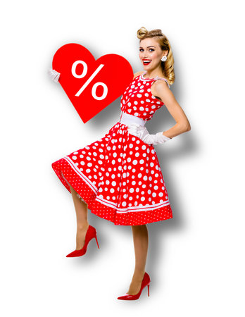Woman holding red paper heart shape with % sign. Full length body portrait of happy pin up girl. Blond model at retro fashion rockabilly sales ad concept. White background. Valentine or like symbol.の写真素材
