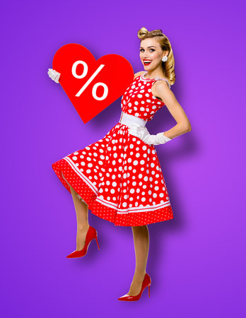 Woman holding red paper heart shape with % sign. Full length body portrait of happy pin up girl. Blond model at retro fashion rockabilly sales ad concept. Purple background. Valentine or like symbol.の写真素材