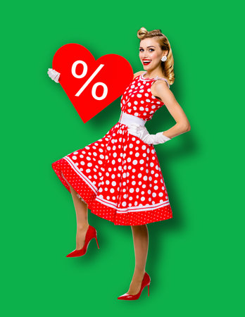 Woman holding red paper heart shape with % sign. Full length body portrait of happy pin up girl. Blond model at retro fashion rockabilly sales ad concept. Green background. Valentine or like symbol.の写真素材
