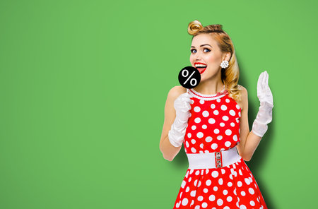 Very tasty discounts, rebates, deals concept image. Cheerful beautiful woman licking signboard with % sign, dressed in pinup red dress, isolated over green background. Black Friday sales ad.の写真素材
