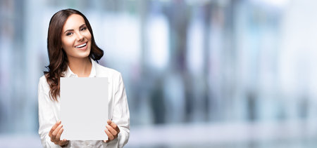 Happy smiling brunette businesswoman showing blank signboard with copy space area for some slogan message, over blured office background. Success in business concept.の写真素材