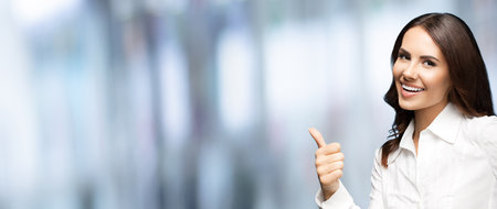 Portrait of very happy smiling young excited businesswoman, showing thumb up hand sign gesture. Success in business concept. Cheerful woman standing on blurred office background.の写真素材