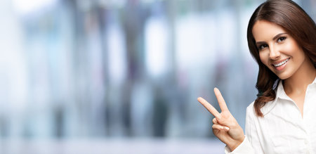 Portrait of happy smiling businesswoman, showing two fingers or victory gesture, with blank copy space for text, over blurred office background. Success in business concept.の写真素材