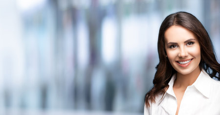Portrait of happy smiling young cheerful brunette businesswoman, with blank copy space area, over blured office background. Success in business concept.の写真素材