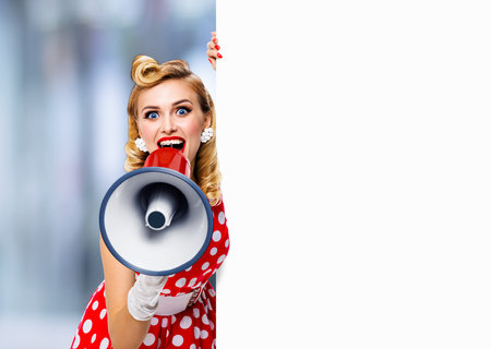 Portrait image of beautiful woman holding megaphone, shout advertising something. Girl in red pin up style dress with mega phone loudspeaker empty banner with copy space. Blurred mall backgroundの写真素材