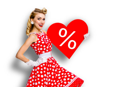 Woman holding red paper heart shape with % sign.  Portrait image of happy pin up girl. Blond model at retro fashion rockabilly sales ad concept. White background. Valentine or like symbol.の写真素材