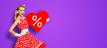 Woman holding red paper heart shape with % sign.  Portrait image of happy pin up girl. Blond model at retro fashion rockabilly sales ad concept. Violet background. Valentine or like symbol.の写真素材