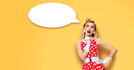 Studio portrait image of thinking woman in red pin up dress. Blond pinup thougthfull girl on yellow background. White empty blank speech sign bubble, having idea.の写真素材