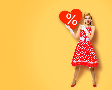 Woman holding red paper heart shape with % sign. Full length body portrait of happy pin up girl showig pointing signboard. Sales ad concept. Yellow background. Valentine or like symbol. Rockabilly.の写真素材