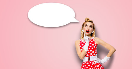 Studio portrait image of thinking woman in red pin up dress. Blond pinup thougthfull girl on rose pink background. White empty blank speech sign bubble, having idea.の写真素材