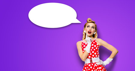 Studio portrait image of thinking woman in red pin up dress. Blond pinup thougthfull girl on violet purple background. White empty blank speech sign bubble, having idea.の写真素材