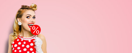 Very tasty discounts, rebates, deals, sales concept. Cheerful beautiful woman licking signboard with % sign, dressed in pinup red dress isolated on pink background. Happy girl at studio. Wide pictureの写真素材