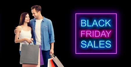 Big deals, discounts offers concept - happy couple with shopping bags, looking at each other. Isolated over dark background. Black friday sales neon light sign text.の写真素材