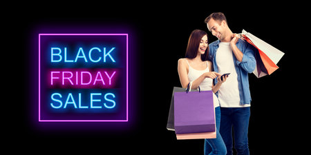 Big deals, discounts offers concept - happy smiling couple with shopping bags, and cellphone. Isolated over dark background. Black friday sales neon light sign text.の写真素材