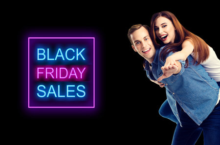 Big deals, discounts offers concept - happy excited couple, in piggyback pose, isolated over dark background. Black friday sales neon light sign text.の写真素材