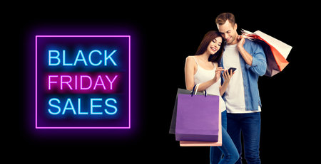 Big deals, discounts offers concept - happy couple holding shopping bags, looking at mobile phone, standing close to each other. Black color background.の写真素材